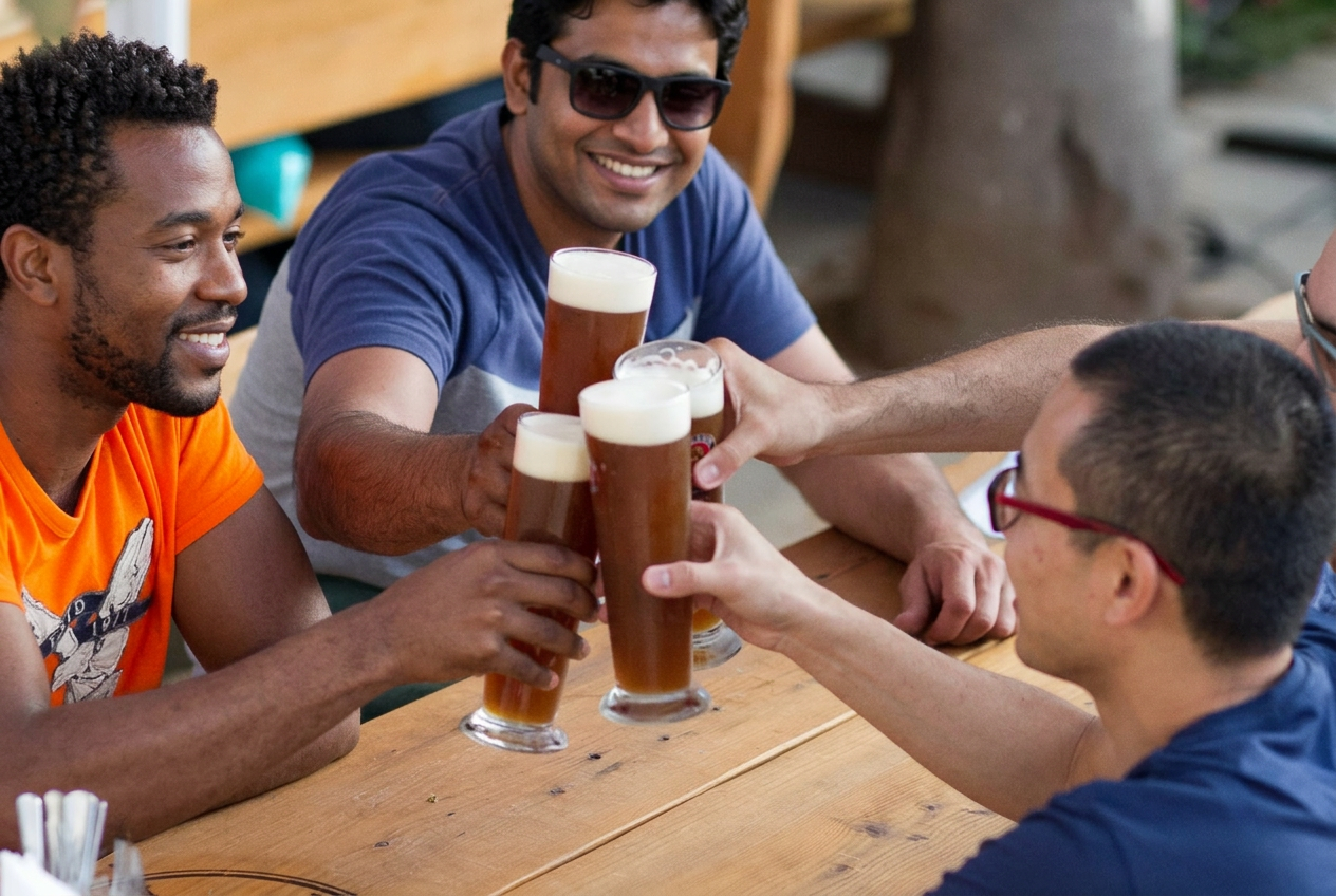 חבר'ה מרימים כוסית בירה Paulaner סביב שולחן עץ — הרגע שבאמת נהנים מהאירוע